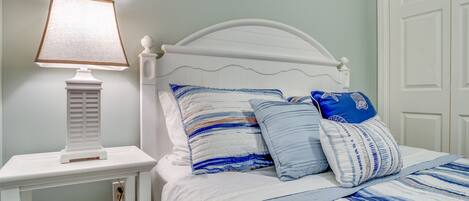 Una cama bien hecha con ropa de cama a rayas, una mesita de noche blanca con una lámpara y un estante con bloques que deletrean "BEACH".