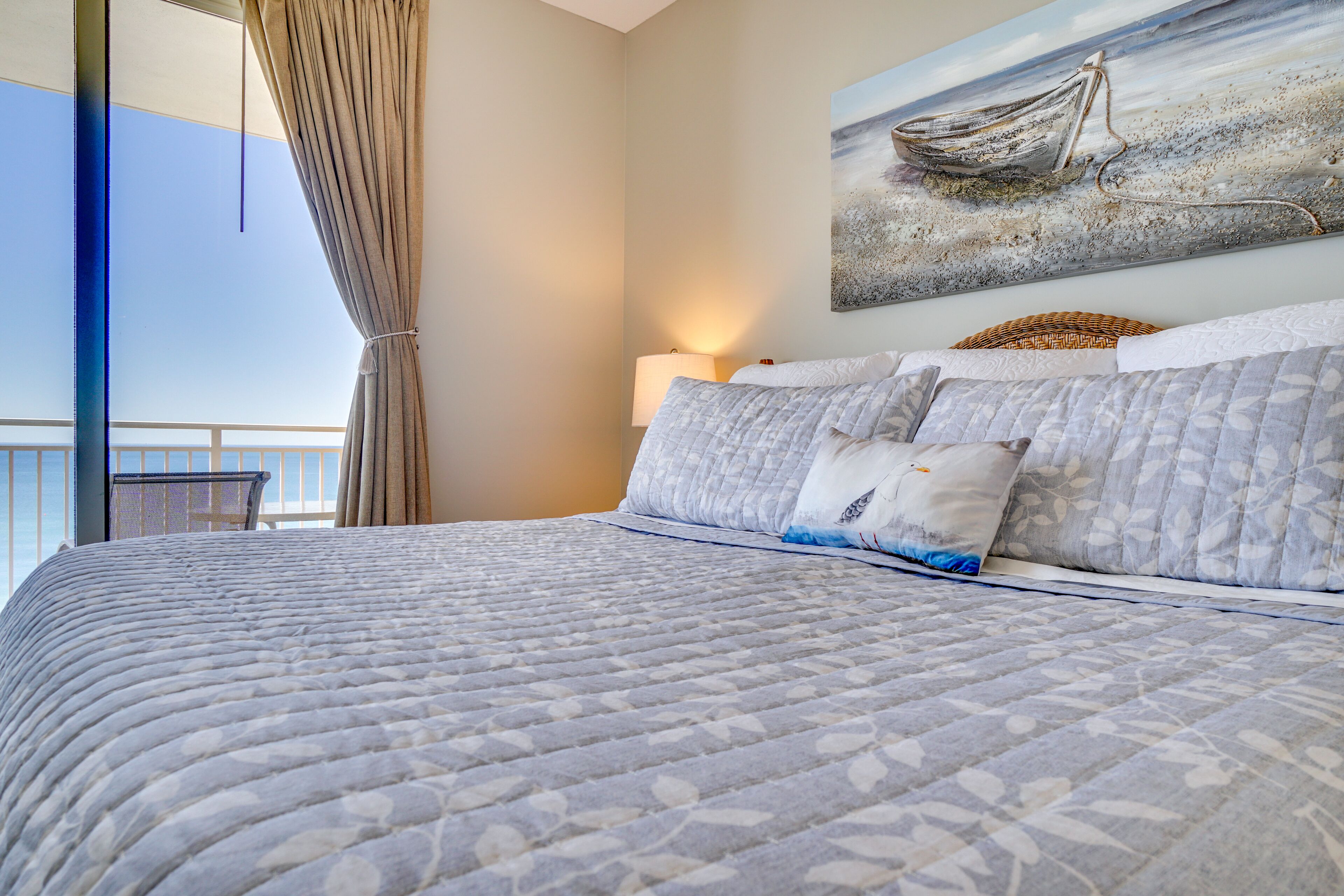Een slaapkamer met een groot bed, uitzicht op zee door een raam, en een schilderij van een boot aan de muur.