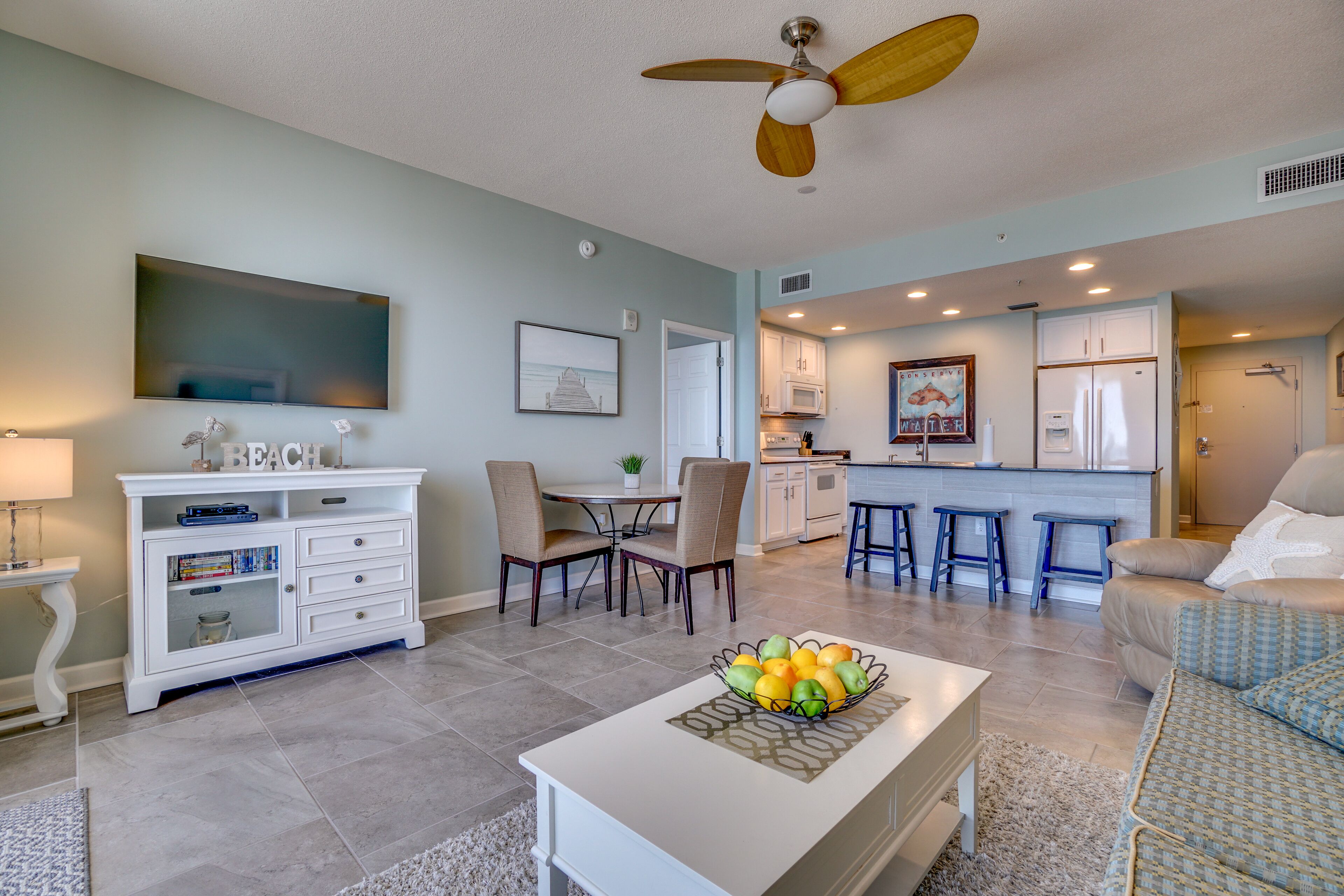 A modern living room with a white coffee table, a dining area with chairs, a kitchen with white cabinets, and a ceiling fan.