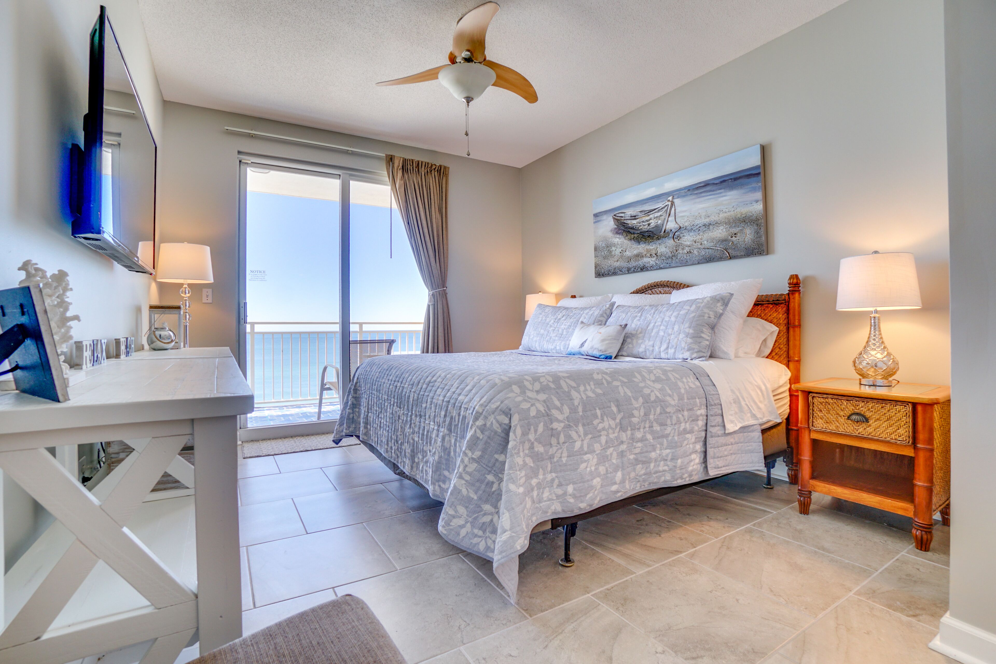 A bedroom with a bed, a desk, a ceiling fan, a painting, and a view of the outside.