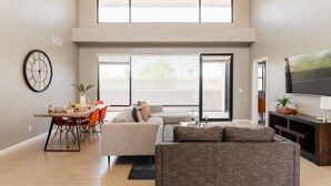 A modern living room with a sofa, dining table, and chairs. There is a large window with blinds, a clock on the wall, and a TV on a stand. - Ivory I By AvantStay | Modern Home w/ Stunning Views | Close To Old Town (Scottsdale)