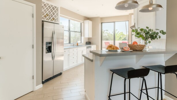 A modern kitchen with white cabinets, a stainless steel refrigerator, a large window, and a central island with bar stools. - Ivory I By AvantStay | Modern Home w/ Stunning Views | Close To Old Town (Scottsdale)