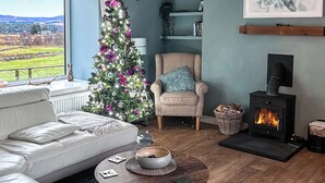 Un salon chaleureux avec un canapé blanc, un sapin de Noël, une cheminée et une vue sur l’extérieur.