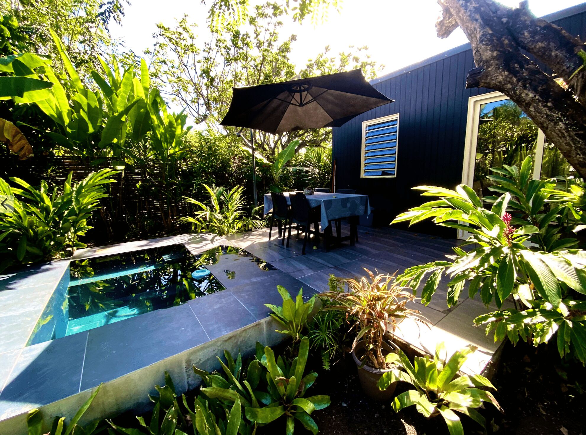 Une piscine dotée d’une terrasse carrelée, entourée d’une végétation luxuriante et agrémentée d’un espace repas avec des chaises et une table placés sous un parasol.