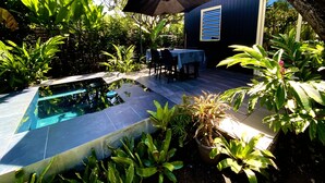 Une piscine dotée d’une terrasse carrelée, entourée d’une végétation luxuriante et agrémentée d’un espace repas avec des chaises et une table placés sous un parasol.