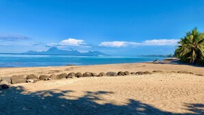 Une plage avec une digue rocheuse, une eau d’un bleu limpide et des montagnes qui se dessinent à l’horizon.