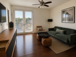 A modern living room with a sofa, a coffee table, and a chair. There is a ceiling fan, a floor lamp, and a large window with blinds.