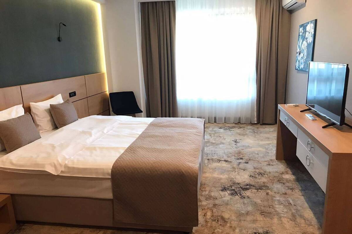 A hotel room with a bed, a desk with a TV, a chair, and a window with curtains.