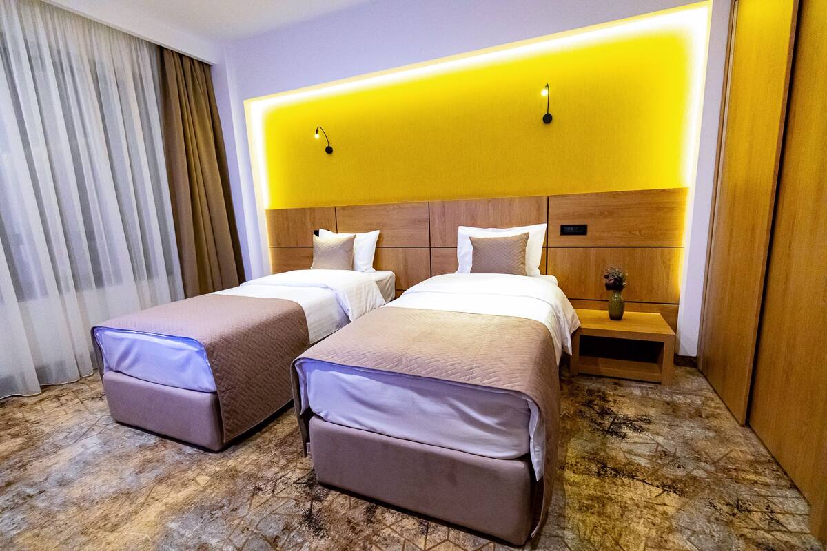 A hotel room with two beds, a wooden headboard, a nightstand with a vase, and a large window with curtains.