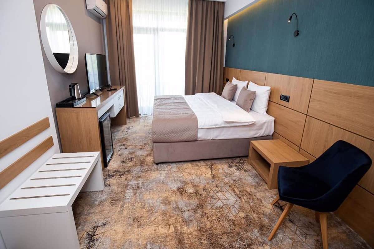 A hotel room with a bed, a desk, a chair, a mirror, and a wall-mounted light.