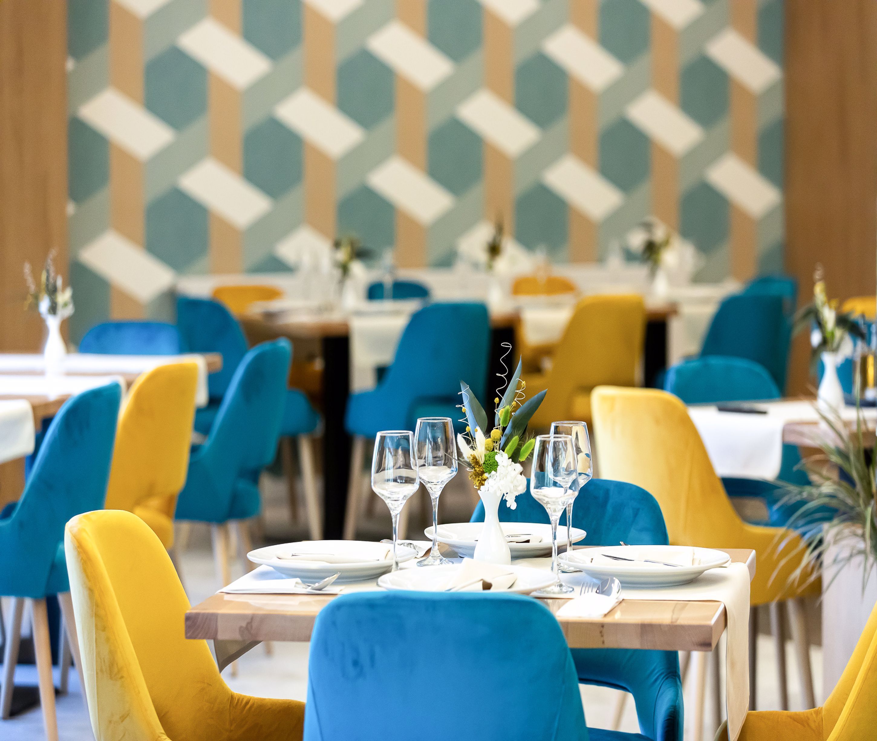 A dining area with tables set for two, featuring blue and yellow chairs, and a patterned wall in the background.