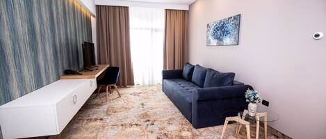 Una habitación de hotel moderna con un sofá azul oscuro, una mesita consola blanca, un escritorio con una silla y una mesa de centro de cristal.