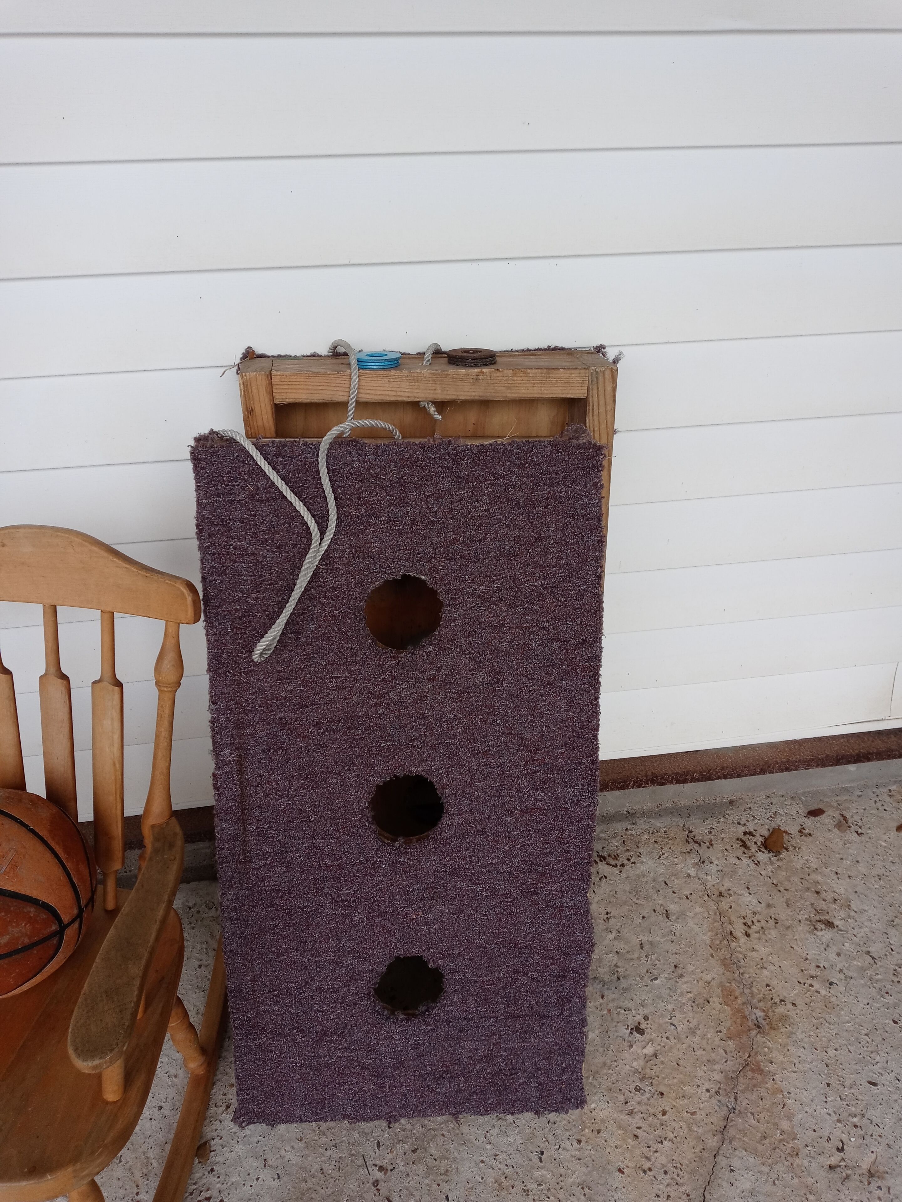 A purple carpeted board with three holes, a wooden chair, and a wooden box against a white wall.