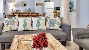 Una sala de estar con un sofá gris, cojines a juego, una mesa de centro de madera con un libro y un objeto decorativo rojo, y una zona de comedor con sillas de madera y una mesa.