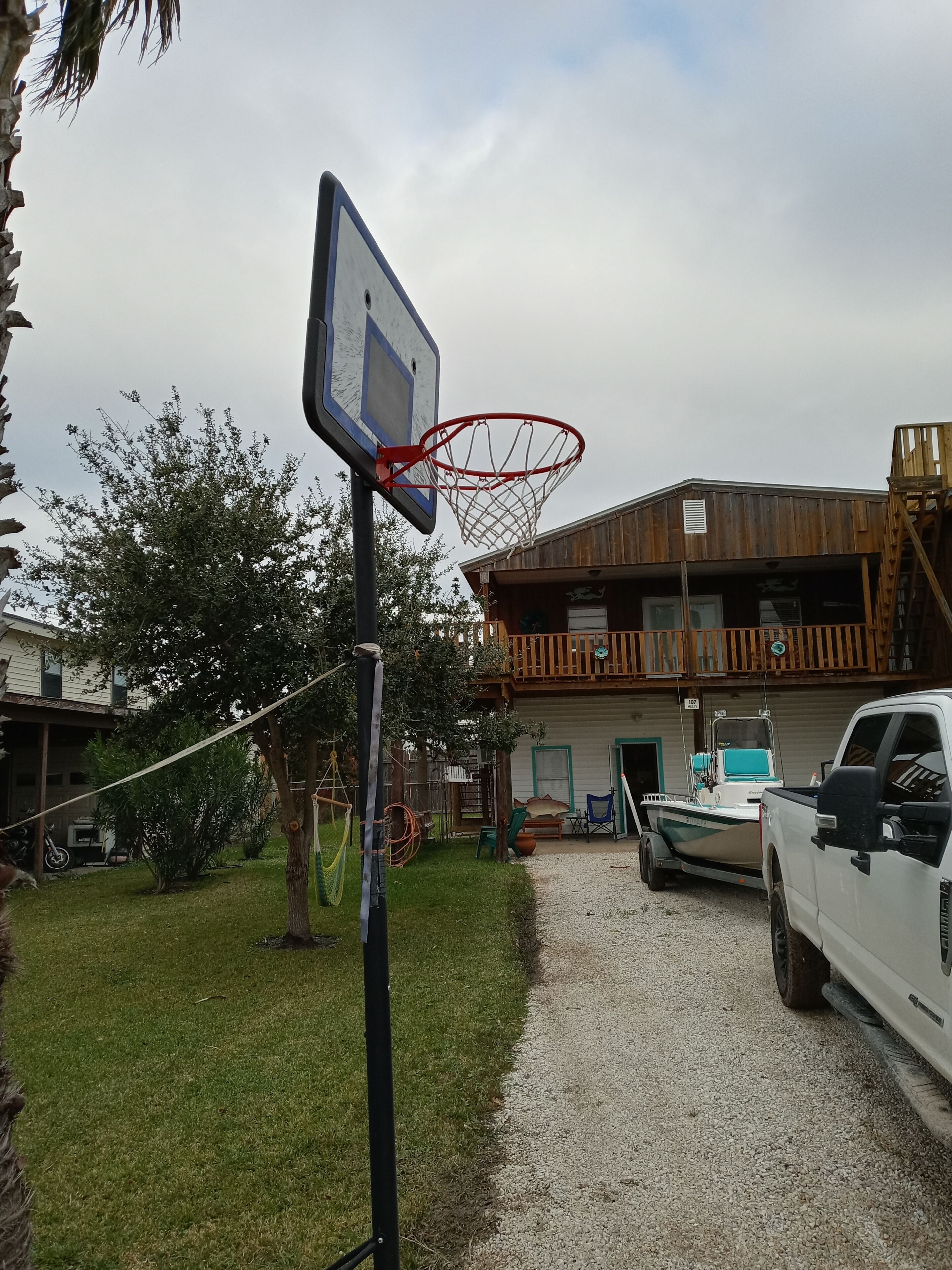 A basketball hoop in a backyard with a gravel path, a white truck, and a wooden building with a balcony.