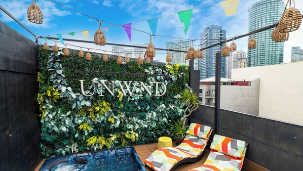 Uma área de estar no terraço com hidromassagem, espreguiçadeiras e uma parede com vegetação, exibindo o texto 'UNWIND'.