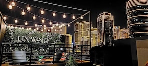 A rooftop bar with string lights, a sign reading 'UNWIND', and a city skyline in the background.