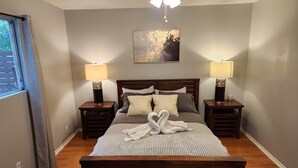 A neatly arranged bedroom with a wooden bed, two bedside tables with lamps, a framed artwork on the wall, and a window with curtains.