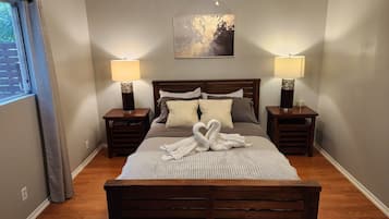 A neatly arranged bedroom with a wooden bed, two bedside tables with lamps, a framed artwork on the wall, and a window with curtains.