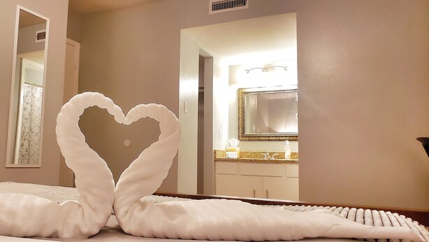 A hotel room with a bed featuring a heart-shaped towel arrangement and a large mirror reflecting the bathroom.