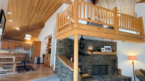A spacious loft bedroom with a stone fireplace, wooden balcony, and a kitchen area visible in the background. - GM239: Private Hot Tub + Family/Dog Friendly + Central AC (Sisters)