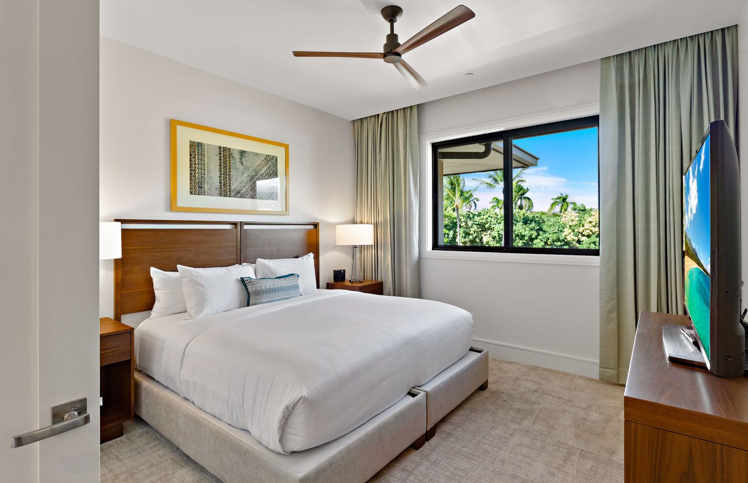 A hotel room with a large bed, a ceiling fan, a TV, and a window with a view of palm trees.
