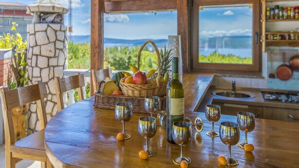 Une table en bois, une bouteille de vin et plusieurs verres, un panier de fruits et un pilier en pierre.