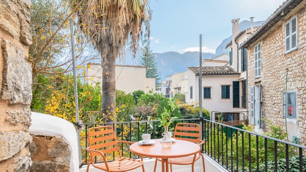 Un balcon avec une table et des chaises, donnant sur une rue bordée d’immeubles et d’arbres.