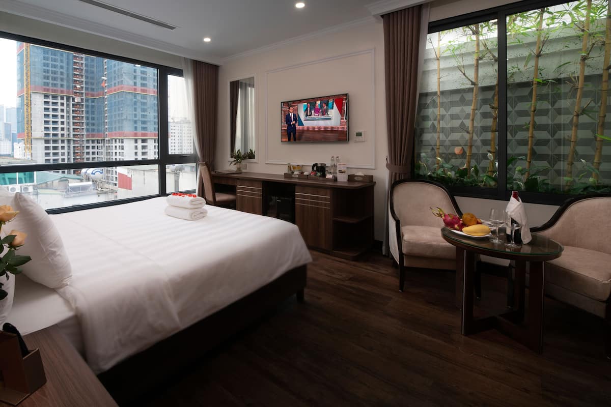 A hotel room with a large bed, a desk, a chair, a small table with fruits and a wine glass, and a TV on the wall.