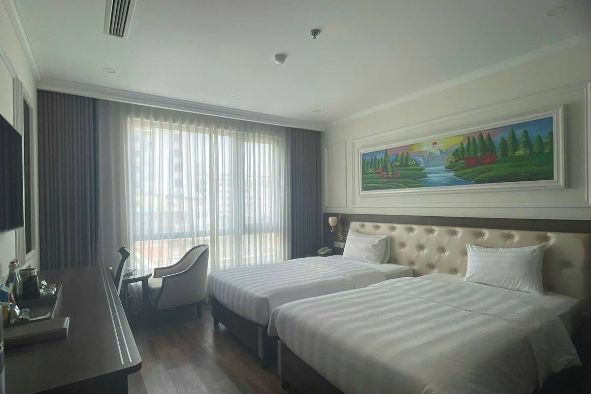 A hotel room with two beds, a desk, a chair, a painting on the wall, and a window with curtains.