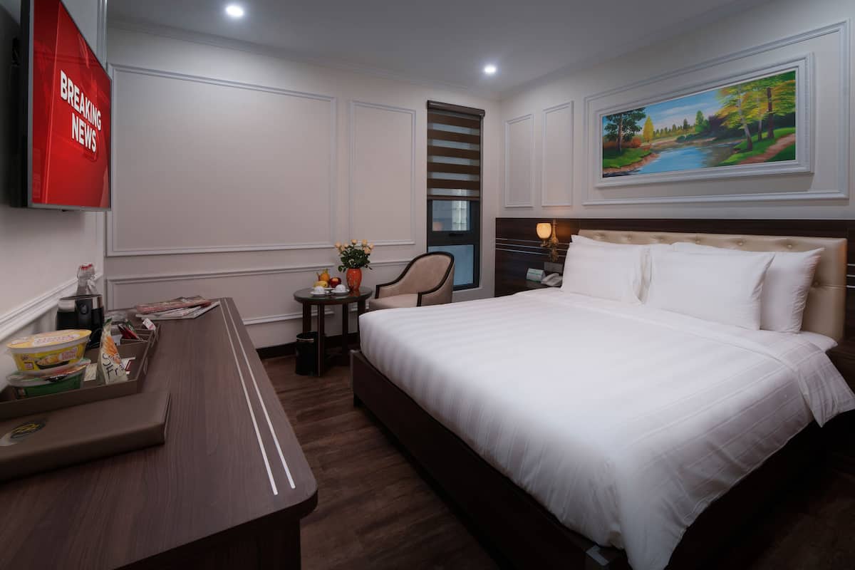 A hotel room with a large bed, a desk with breakfast items, a painting on the wall, and a window with blinds.