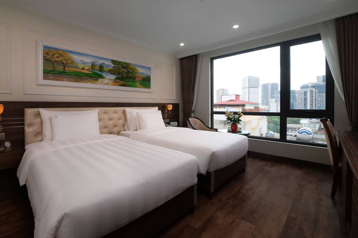 A hotel room with two beds, a painting on the wall, a window with a city view, and a vase with flowers on the windowsill.