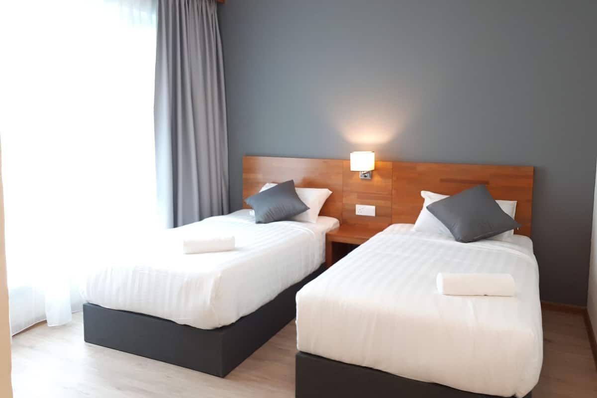 A hotel room with two single beds, a wooden headboard, a nightstand with a lamp, and a large window with curtains.