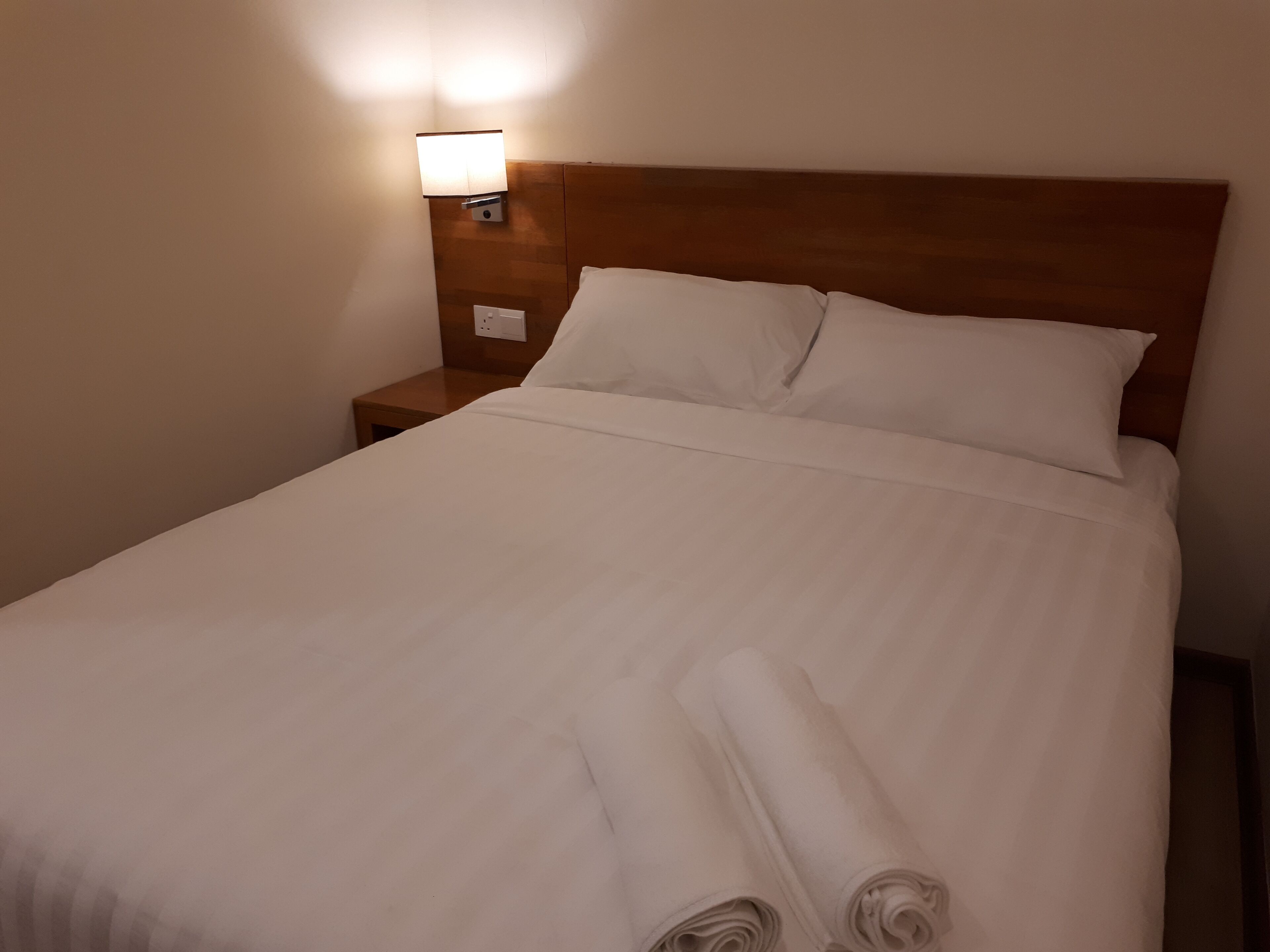 A neatly made bed with white linens and pillows, a wooden headboard, and a bedside table with a lamp.