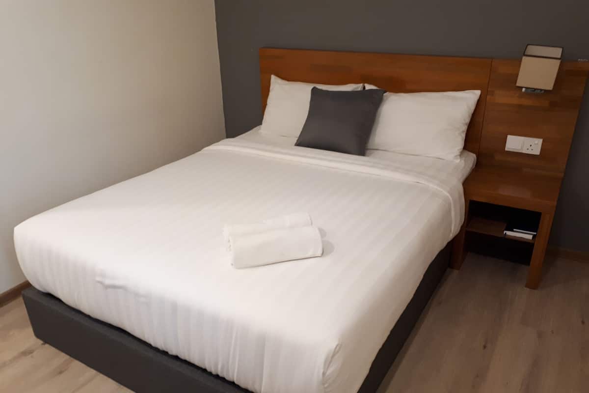 A neatly made bed with white linens and a folded towel, a wooden headboard, a nightstand with a lamp, and a wooden floor.