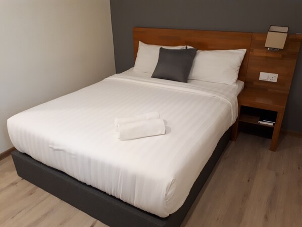 A neatly made bed with white linens and a folded towel, a wooden headboard, a nightstand with a lamp, and a wooden floor.