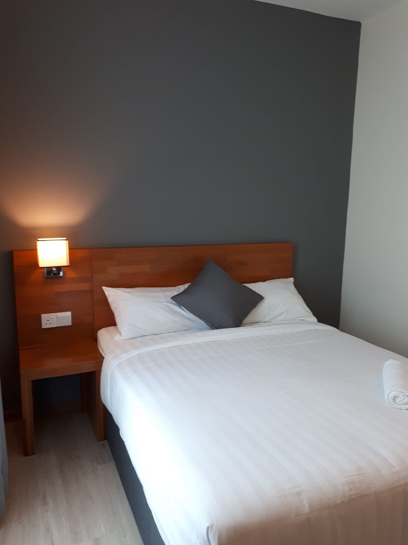 Un lit bien fait, avec du linge de lit blanc et un oreiller gris, une tête de lit en bois, une lampe de chevet et une prise électrique sur la table de chevet.