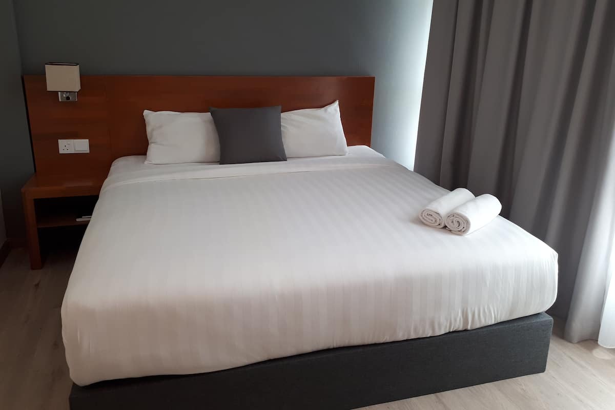 A neatly made bed with white linens and rolled towels, a wooden headboard, a bedside table with a lamp, and a power outlet.