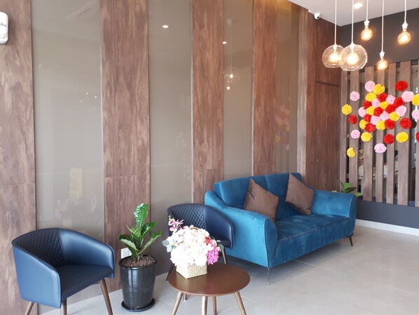 Un área de recepción moderna con un sofá azul, dos sillones, una planta en maceta y una pared decorativa con flores de colores.
