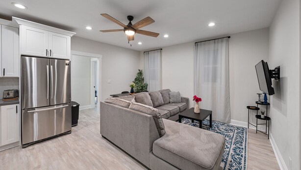 A modern living space with a kitchen, a grey sectional sofa, a ceiling fan, and a flat-screen TV mounted on the wall.