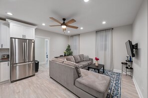 A modern living space with a kitchen, a grey sectional sofa, a ceiling fan, and a flat-screen TV mounted on the wall.