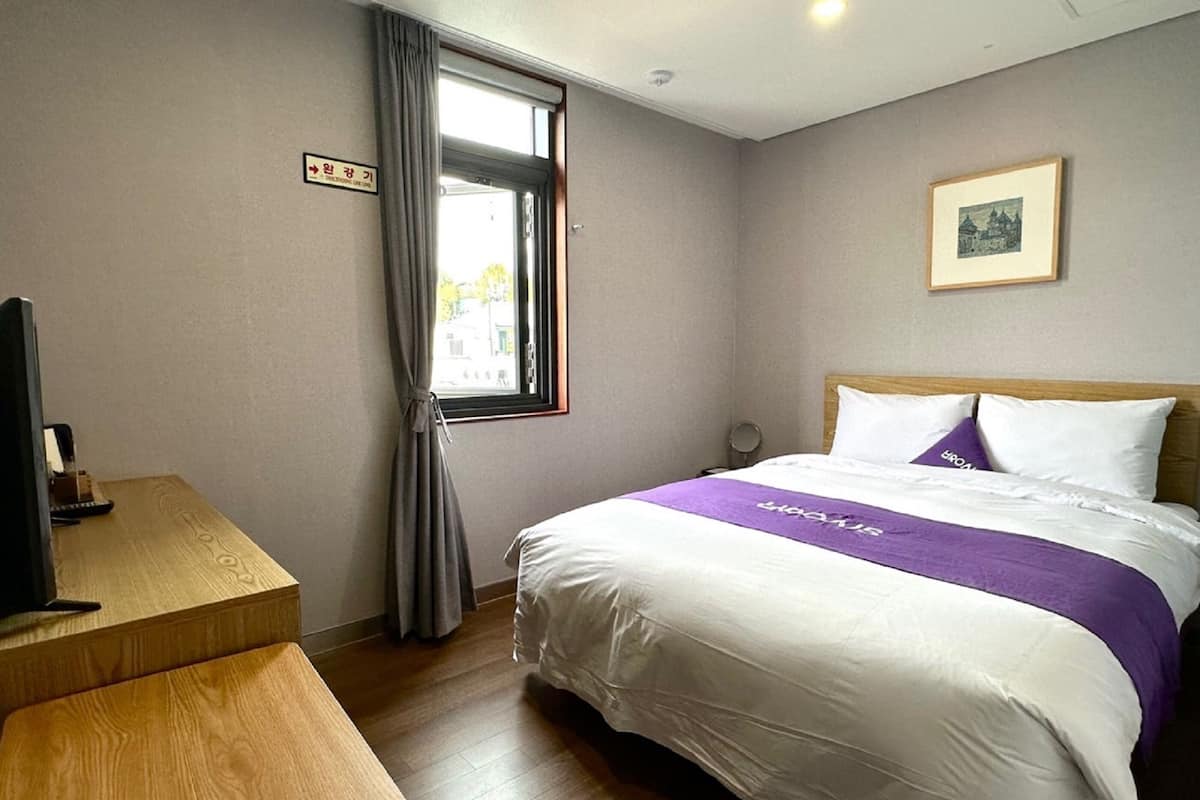 Habitación de hotel con cama, escritorio, ventana con cortinas, un cuadro enmarcado en la pared y un cartel que indica el número de habitación.