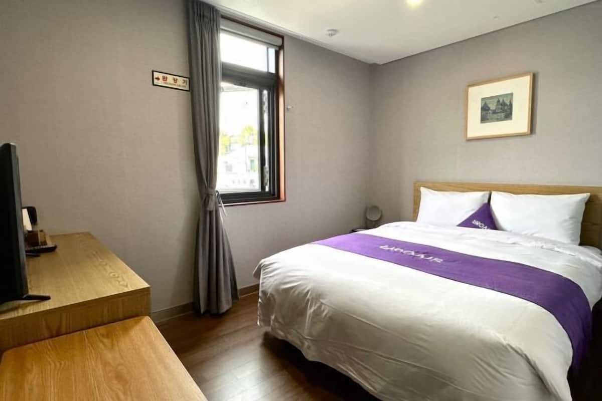 Habitación de hotel con cama, escritorio, televisor, ventana con cortinas e imagen enmarcada en la pared.