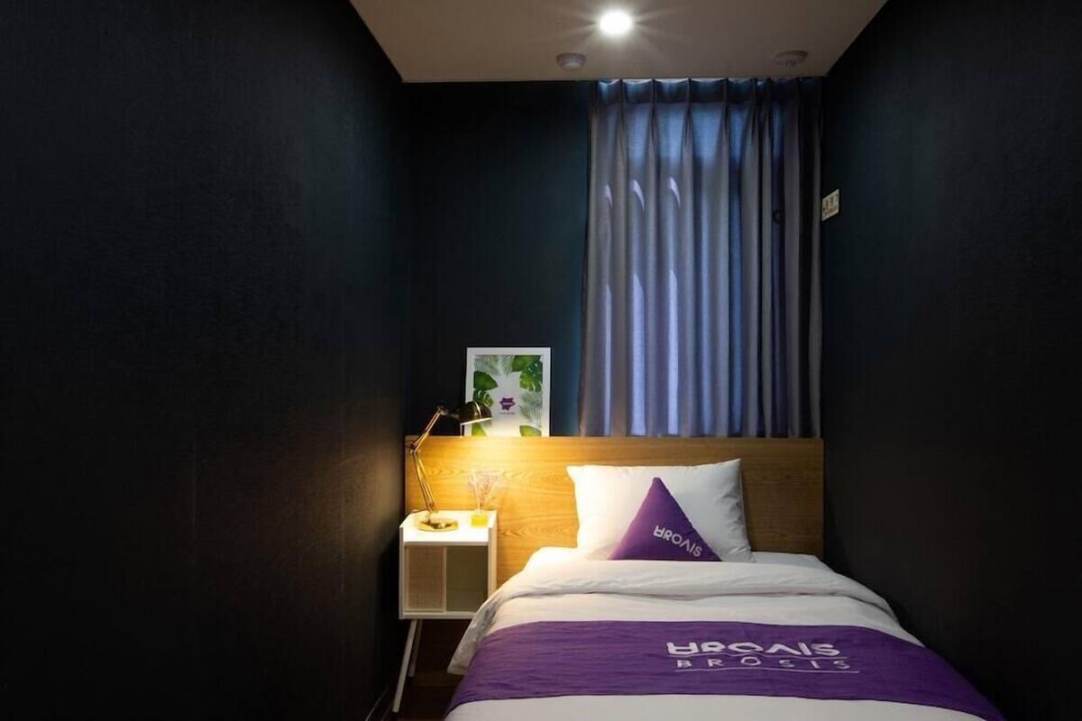 Una habitación de hotel con una cama, una mesita de noche, una lámpara, un cuadro enmarcado y una ventana con cortinas.