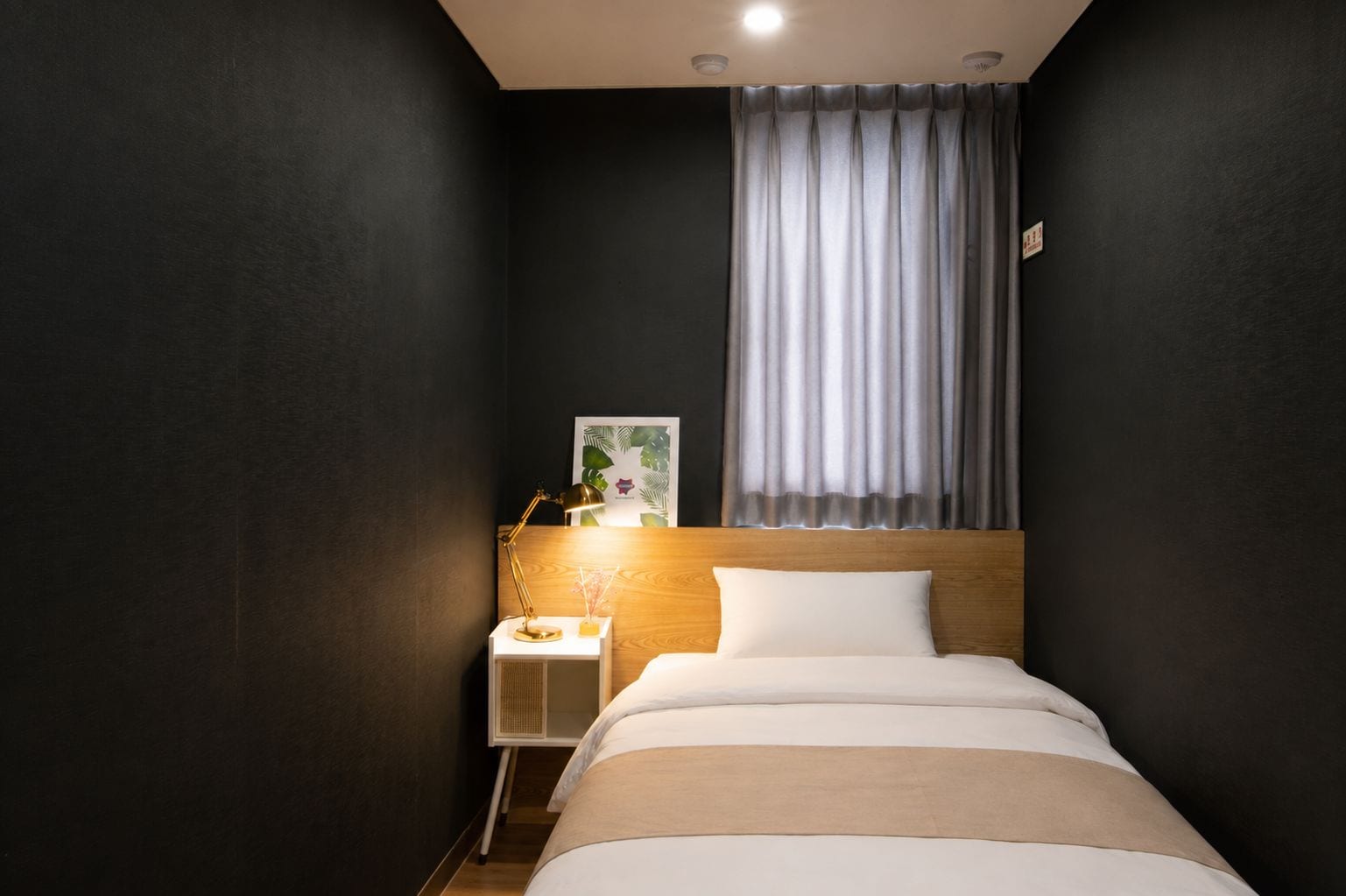 A hotel room with a bed, a nightstand, a lamp, a window with curtains, and a wall-mounted picture.