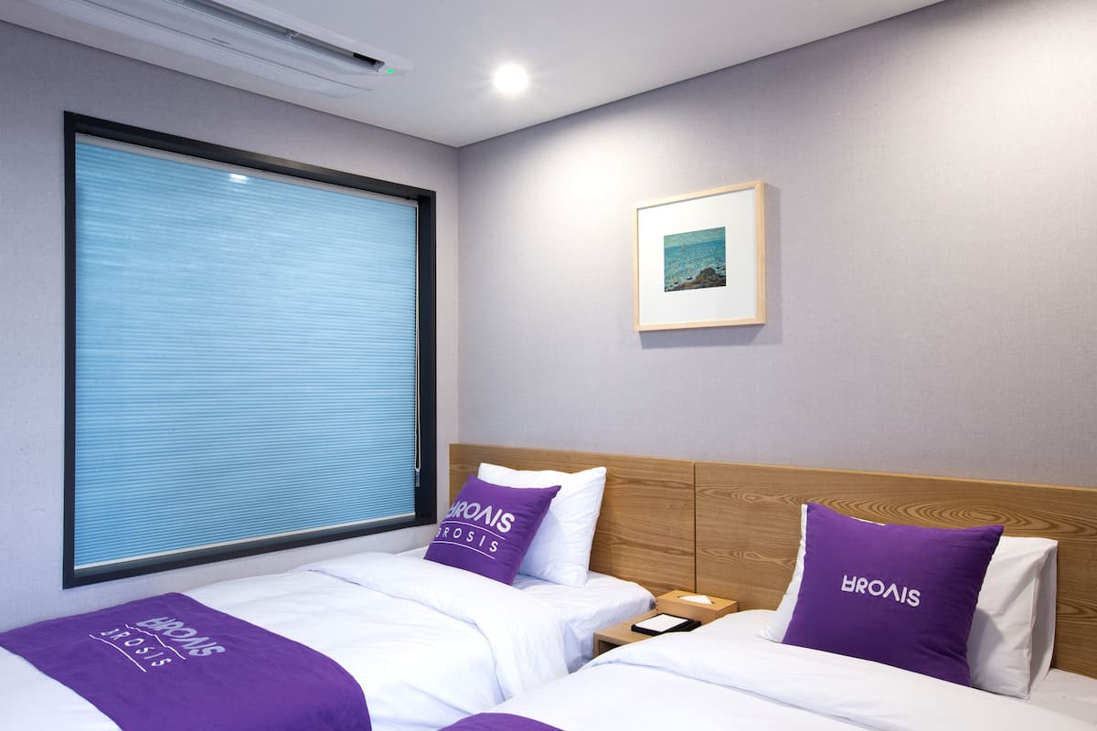 Habitación de hotel con dos camas, un ventanal con persianas, un cuadro enmarcado en la pared y un cabecero de madera.