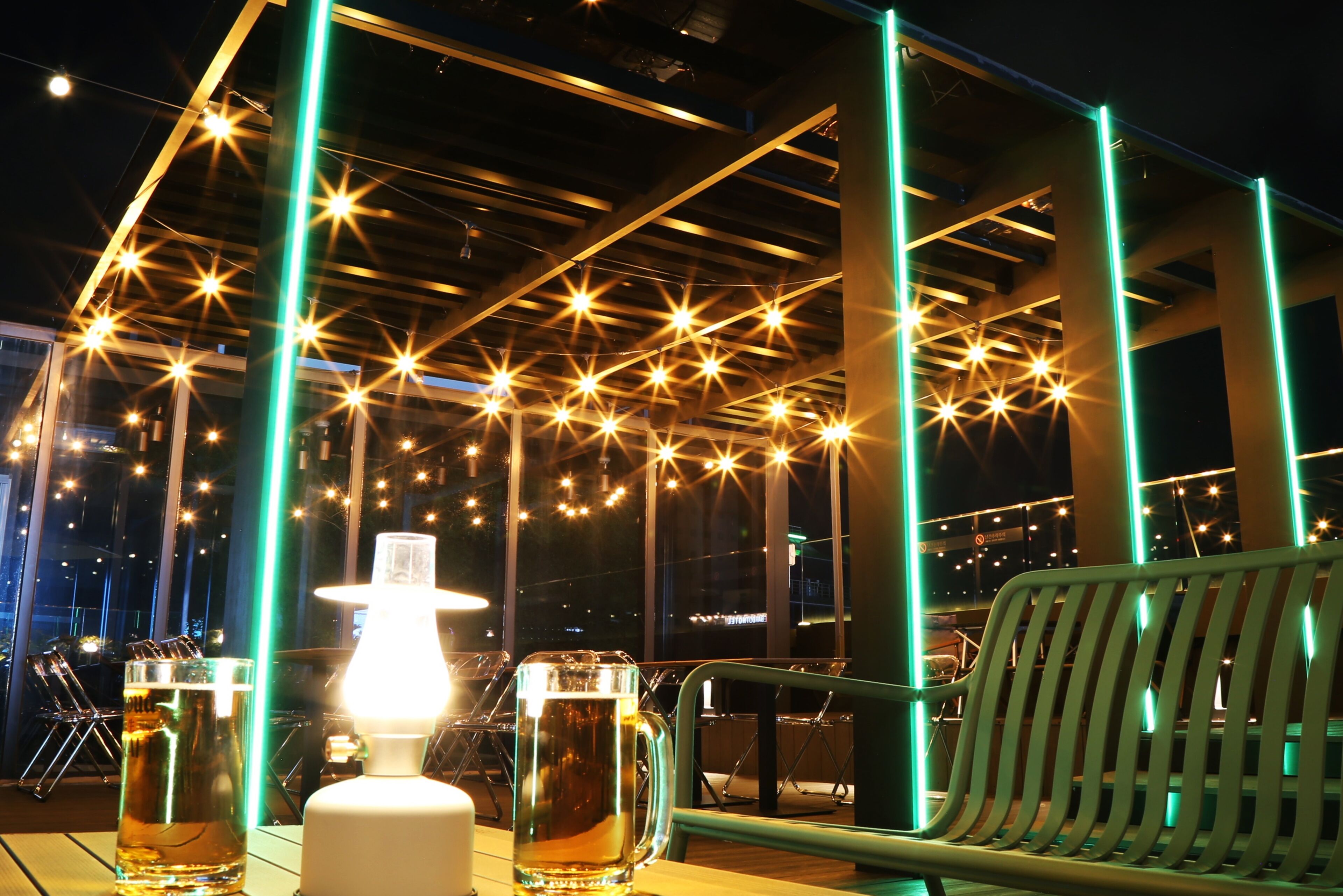 A table with two glasses of beer and a lit lamp, set against a backdrop of a metal structure with star-shaped lights.