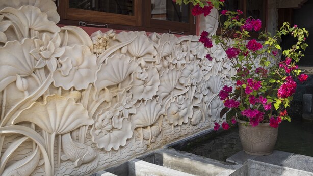 A detailed stone carving with floral and fish motifs, a potted plant with pink flowers, and a window with wooden frames. - Kemangi Apartments and Yoga Space (Ubud)