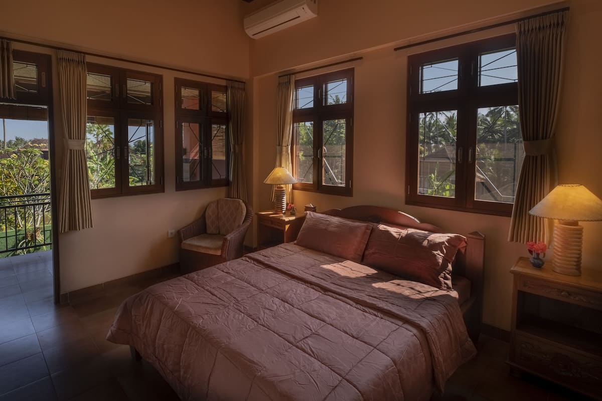 A bedroom with a large bed, two bedside tables with lamps, a chair, and multiple windows with curtains.