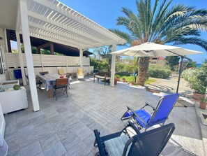 Une terrasse extérieure dotée d’un espace salon couvert, d’une table avec des chaises et de plantes en pot.